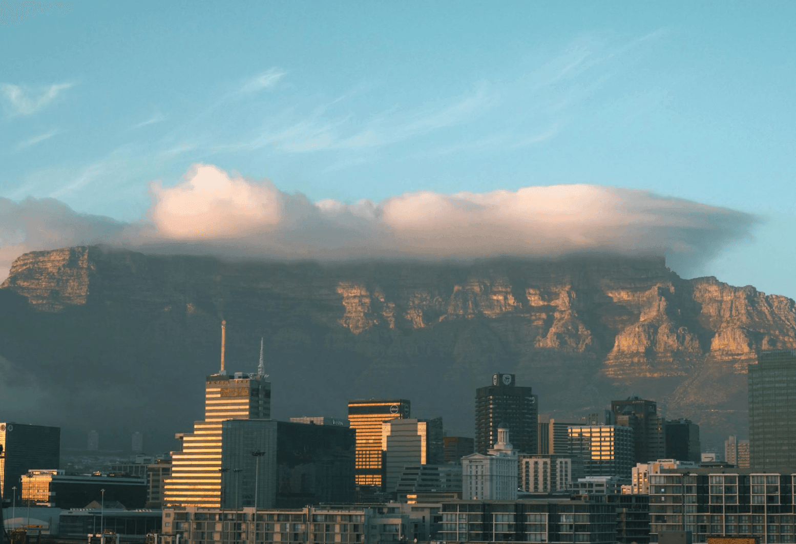 Cloud-covered mountain towering over a city skyline with modern buildings under a clear blue sky.